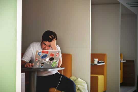 How to Counter Workplace Burnout: a man in white shirt with his hand on his forehead using mac book pro